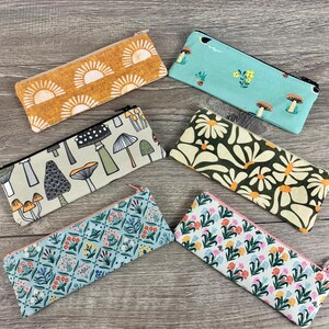 May include: Six different fabric pencil cases with various patterns. The patterns include mushrooms, flowers, and a sunburst design. The pencil cases are all rectangular and have a zipper closure.
