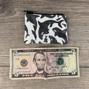 Puede incluir: Una peque&ntilde;a bolsa blanca con cremallera con un estampado en blanco y negro de orcas. La bolsa est&aacute; sobre una superficie de madera junto a un billete de cinco euros.