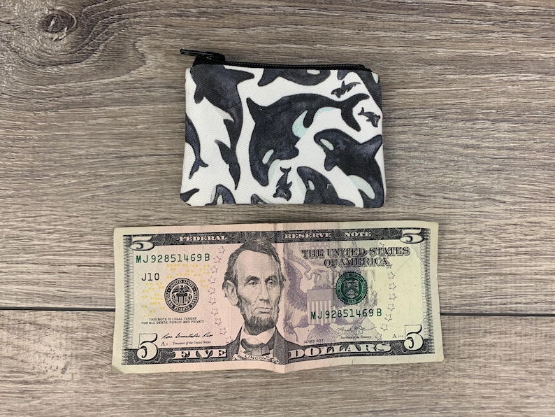 Puede incluir: Una peque&ntilde;a bolsa blanca con cremallera con un estampado en blanco y negro de orcas. La bolsa est&aacute; sobre una superficie de madera junto a un billete de cinco euros.