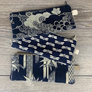 May include: Three navy blue zippered pouches with white patterns. The top pouch features floral and cloud designs, the middle one has a dragonfly pattern, and the bottom one showcases bamboo and geometric motifs. Each pouch has a small fabric loop.