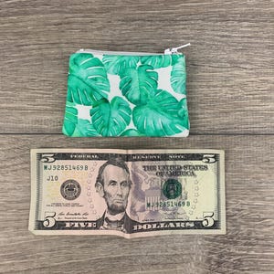 Puede incluir: Una peque&ntilde;a cartera de monedas verde y blanca con cierre de cremallera. La cartera tiene un estampado de hojas tropicales. La cartera est&aacute; sobre una superficie de madera junto a un billete de cinco euros.