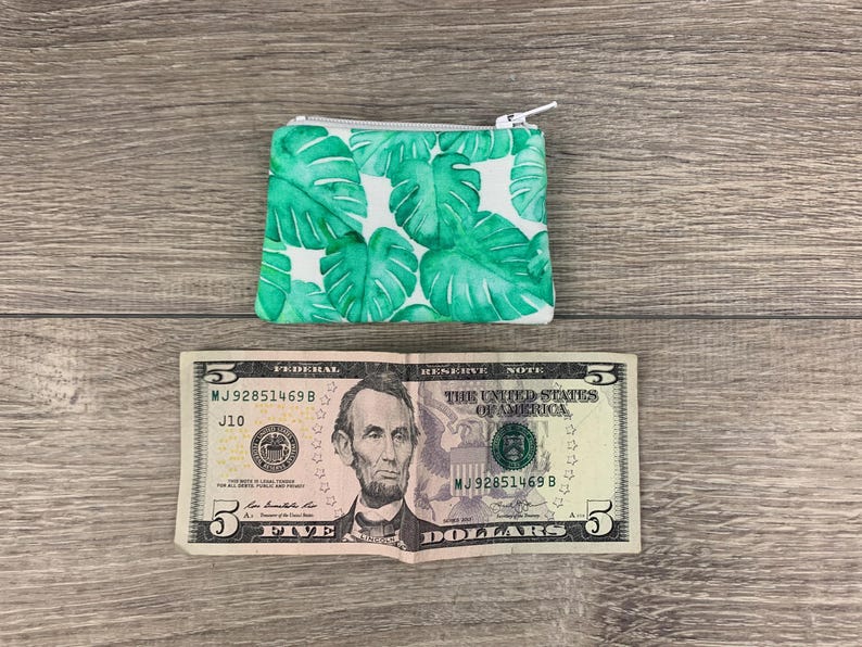 Puede incluir: Una peque&ntilde;a cartera de monedas verde y blanca con cierre de cremallera. La cartera tiene un estampado de hojas tropicales. La cartera est&aacute; sobre una superficie de madera junto a un billete de cinco euros.