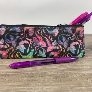 May include: A black pencil case with a zipper closure. The case is decorated with a colorful watercolor pattern of dragons. A purple pen is lying in front of the case.