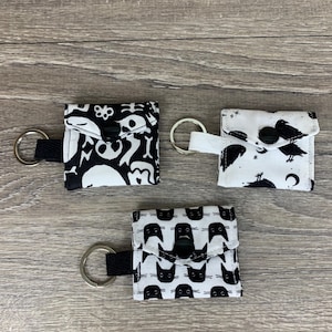 May include: Three small, square fabric pouches with key rings. Each pouch features a different black and white pattern: skulls, crows, and cat faces. The pouches have a flap closure with a black button and a black or white fabric loop for the key ring.