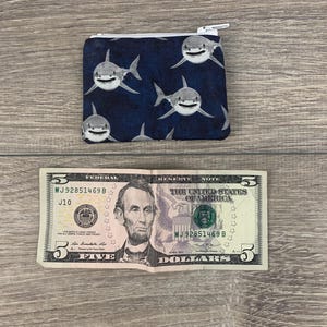 Puede incluir: Una peque&ntilde;a bolsa con cremallera con fondo azul y un patr&oacute;n de tibur&oacute;n blanco. La bolsa est&aacute; sobre una superficie de madera junto a un billete de cinco euros.