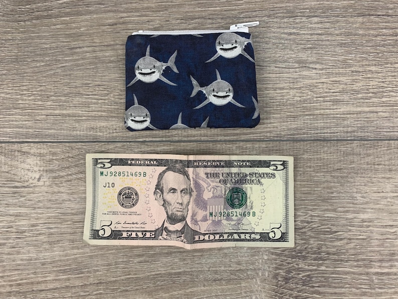 Puede incluir: Una peque&ntilde;a bolsa con cremallera con fondo azul y un patr&oacute;n de tibur&oacute;n blanco. La bolsa est&aacute; sobre una superficie de madera junto a un billete de cinco euros.