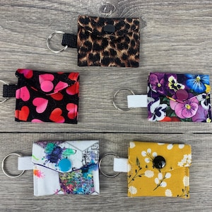 May include: Five different keychains with fabric pouches. The pouches are decorated with various patterns, including hearts, flowers, and animal print. Each keychain has a metal ring and a white cord.