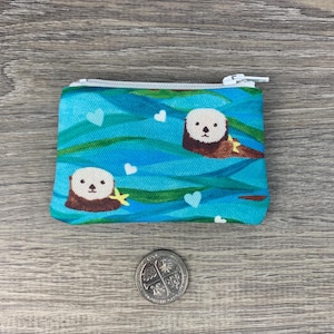 Puede incluir: Una bolsa azul con cremallera blanca. La bolsa tiene un patr&oacute;n de nutrias marinas nadando en agua azul con corazones blancos.