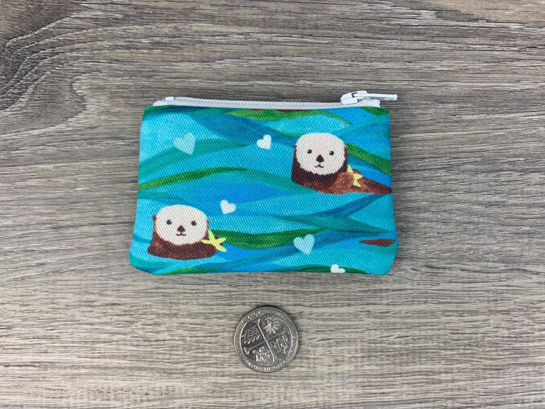 Puede incluir: Una bolsa azul con cremallera blanca. La bolsa tiene un patr&oacute;n de nutrias marinas nadando en agua azul con corazones blancos.