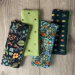 Könnte beinhalten: Fünf rechteckige Reißverschlusstaschen in verschiedenen Mustern und Farben. Eine ist einfarbig hellgrün mit gleichmäßig verteilten Löchern. Andere zeigen Blumen- und Insektenmuster in Dunkelgrün, Schwarz und Blau. Jede hat einen kontrastierenden Reißverschluss.