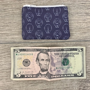 Puede incluir: Una peque&ntilde;a bolsa con cremallera de fondo azul oscuro y un patr&oacute;n blanco repetido de hipop&oacute;tamos. La bolsa est&aacute; sobre una superficie de madera junto a un billete de cinco euros.