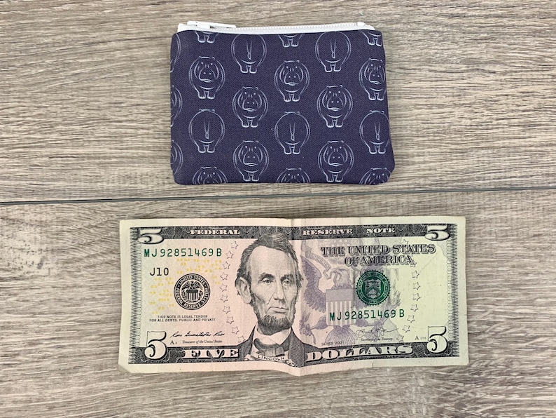 Puede incluir: Una peque&ntilde;a bolsa con cremallera de fondo azul oscuro y un patr&oacute;n blanco repetido de hipop&oacute;tamos. La bolsa est&aacute; sobre una superficie de madera junto a un billete de cinco euros.