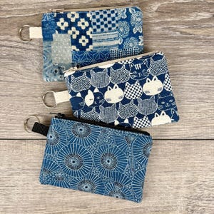 May include: Three small, zippered coin purses with key rings. The purses feature blue and white patterns, including patchwork, cat motifs, and a radial design. Each purse has a different pattern and a contrasting zipper.