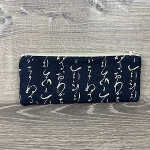 May include: A navy blue pencil case with a white zipper and a repeating white script pattern. The rectangular case is made of fabric and rests on a wooden surface.