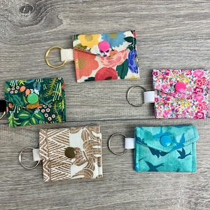 May include: Five keychains with different fabric patterns. Each keychain has a small pocket and a metal ring. The patterns include floral, geometric, and abstract designs. The keychains are a mix of green, blue, pink, and brown.