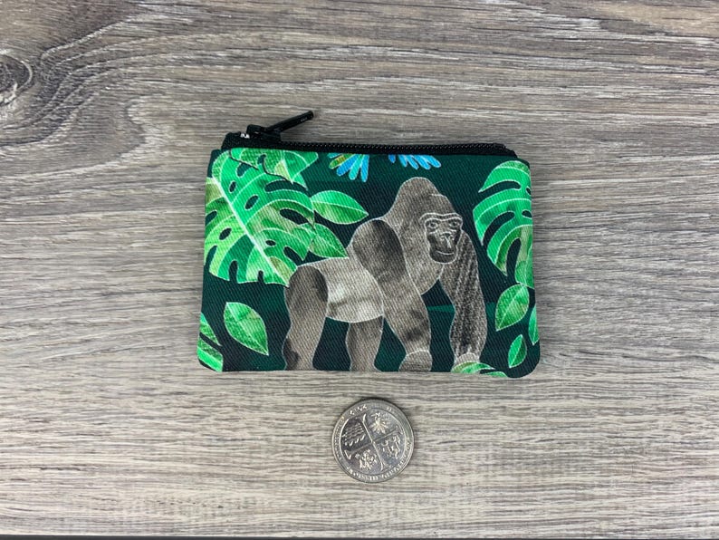 Puede incluir: Una peque&ntilde;a bolsa con cremallera verde y negra con un gorila y hojas tropicales impresas.