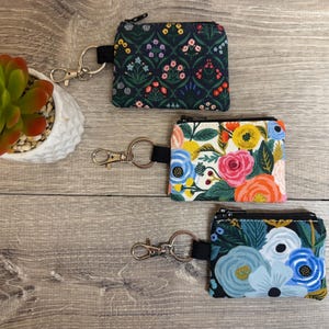 May include: Three rectangular fabric coin pouches with black zippers and metal lobster claw keychains. The top pouch features a dark green background with small floral patterns. The middle pouch has a white background with large colorful roses. The bottom pouch displays blue and white flowers on a dark background.