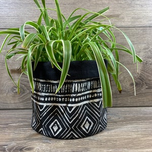 May include: A cylindrical black and white patterned fabric planter pot holding a green and white spider plant. The pot features a geometric design with diamonds and lines. The top edge of the pot is solid black.