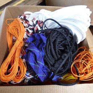 May include: A cardboard box filled with various ropes and cords. The assortment includes orange, black, white, and blue ropes, along with some with red and black accents. The ropes are tightly coiled and bundled.