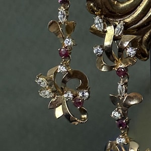 May include: A pair of gold drop earrings featuring a floral-inspired design with curved metal petals, accented by small round red rubies and clear marquise-cut diamonds arranged in a vertical, dangling pattern that catches the light.