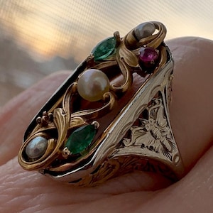 May include: A gold and silver ring with a delicate filigree design. The ring features a cluster of green emeralds, red rubies, and white pearls.
