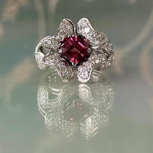 May include: A floral-shaped ring with a square, deep pink gemstone center. The ring is surrounded by smaller, clear gemstones and has a silver-colored band. The ring's design is reflected on the surface below.