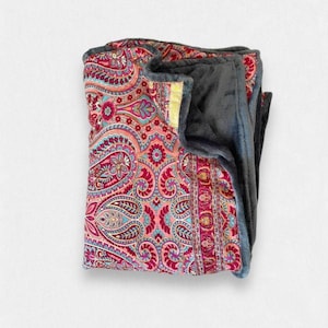 Op de afbeelding: Een gevouwen deken met een paisley-patroon in tinten roze, blauw en goud. De deken heeft een zachte, grijze fleecevoering.
