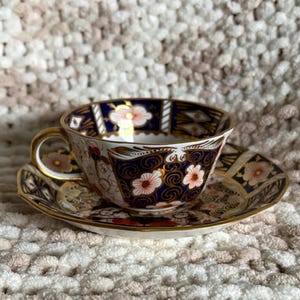 May include: A porcelain teacup and saucer set featuring a deep navy blue background with intricate gold scrollwork and white floral patterns with orange centres. The cup has a gold-trimmed handle and rim, matching the decorative gold-edged saucer.