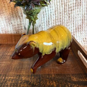 Peut inclure: Une figurine d'ours en céramique avec une finition brillante, présentant un corps inférieur brun foncé et un glaçage jaune moutarde vif sur le dos et la tête. L'ours est représenté en position de marche, la tête baissée, avec une surface lisse.