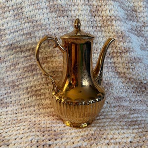 Peut inclure: Une cafetière en métal doré avec une finition réfléchissante, dotée d'un corps cylindrique haut, d'une anse incurvée, d'un long bec verseur et d'un couvercle amovible avec un fleuron pointu. La base est ornée de cannelures verticales.