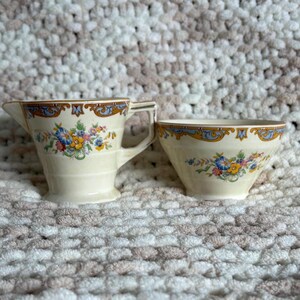 May include: A cream-colored ceramic creamer and sugar bowl set featuring a floral pattern with blue, yellow, and pink flowers. The rim of each piece is decorated with a blue and gold scrollwork border. Both items have a flared shape and a smooth finish.