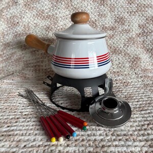 May include: A white enamel fondue pot with a wooden handle and lid knob, featuring red and blue horizontal stripes. It includes a black metal stand, a silver burner, and six metal fondue forks with multi-colored tips in yellow, white, black, blue, green, and orange.