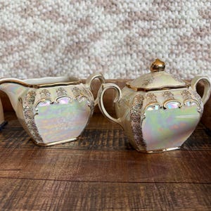 May include: A cream-colored creamer and a lidded sugar bowl with an iridescent finish. Both pieces feature gold-toned ornate filigree patterns along the edges and handles, with a smooth, glossy surface that reflects light in soft pastel hues.