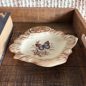 May include: A cream-coloured ceramic trinket dish with a textured brown wood-grain border. The centre features a colourful butterfly illustration perched on floral branches. A small rectangular plaque at the top edge contains embossed text.