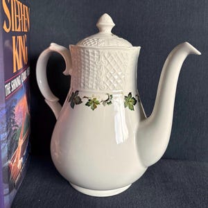 May include: A white ceramic teapot featuring a textured basket-weave pattern on the upper section and lid, accented by a band of green and brown maple leaf illustrations around the middle, complete with a curved handle and a long spout.