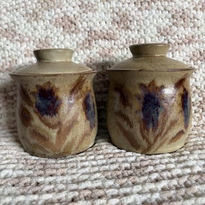 May include: Two matching ceramic jars with lids, featuring a speckled beige glaze and hand-painted floral motifs in shades of deep purple and brown. Each jar has a rounded body, a flared rim, and a fitted lid with a small central knob handle.