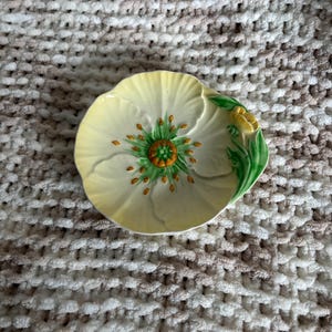 May include: A ceramic dish shaped like a pale yellow flower with a green centre, orange stamens, and green leaves on the right side. The petals have a textured, scalloped edge, and the surface features a glossy finish with detailed relief work.
