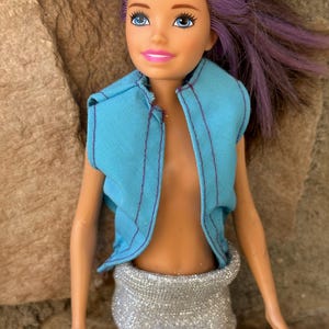 May include: A doll with purple hair wearing a light blue sleeveless vest and a silver sparkly skirt.