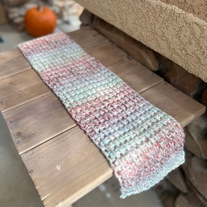 May include: A multi-coloured crocheted scarf with shades of pink, blue, and white. The scarf is lying on a wooden table.