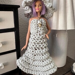 May include: A doll wearing a handmade gray crochet dress and matching wide-brimmed hat. The doll has purple hair and a gold necklace with a charm. The dress is sleeveless and flares out at the skirt. The hat is large and floppy.