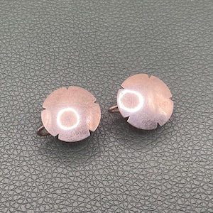 May include: A pair of round, metallic copper-toned clip-on earrings. Each earring features a smooth, slightly domed circular surface with four small, evenly spaced notches along the outer edge, creating a subtle four-petaled flower shape.