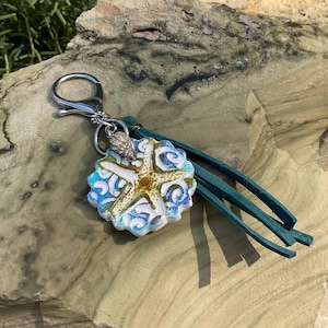 May include: A starfish-shaped keychain with a silver clasp and teal leather tassel. The starfish is white, blue, and gold, with a small silver seashell charm. The keychain is a beach-themed accessory.