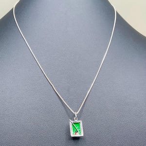 May include: A silver-toned necklace featuring a rectangular pendant shaped like a miniature pool table. The pendant has a bright green surface with a small white cue stick and three tiny billiard balls in red, white, and black colors.