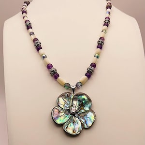 May include: A handcrafted necklace featuring a large, iridescent flower pendant with a central crystal. The necklace has a beaded strand with white, purple, and silver accents. The pendant is approximately 3 inches in diameter.