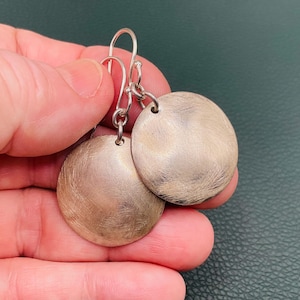 May include: A pair of silver-toned earrings featuring circular, flat discs with a brushed, textured finish. Each disc is suspended from a simple metal hook wire, creating a dangling design that catches the light across the metallic surface.