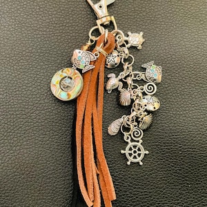 May include: A silver keychain with a lobster clasp, adorned with sea-themed charms. The charms include fish, turtles, seashells, and a ship's wheel. A brown suede tassel hangs below the charms, adding texture and movement to the accessory.