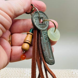 May include: A keychain featuring a weathered, dark green metal key-shaped charm with a square cutout, a light green translucent stone ring, and a brown leather tassel. A wooden bead charm with a small orange sphere is attached to the metal ring.