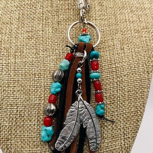 May include: A keychain with a silver clasp and ring, adorned with turquoise, red, and silver beads. Brown, black, and silver suede leather strips hang below the beads. A silver feather charm dangles from the keychain.