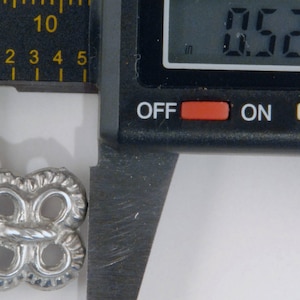 May include: A silver metal button with a decorative design resembling a four-leaf clover. The button is measured with a caliper, showing a measurement of 0.52 inches.