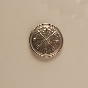 May include: A silver-colored metal coin with a circular design featuring a starburst pattern with eight points.